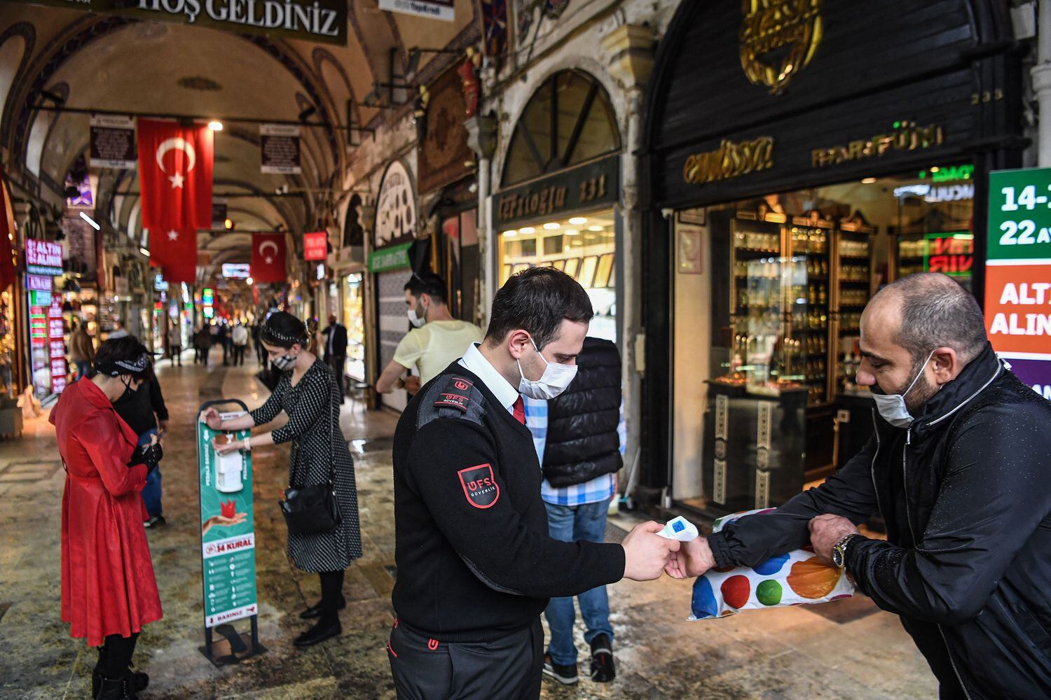 Oficial de segurança verifica a temperatura de um visitante, enquanto mulheres limpam as mãos com gel desinfetante na entrada do icônico Grande Bazar, em Istambul, que reabriu nesta segunda-feira.
