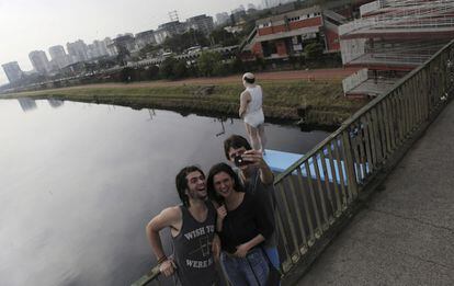 Jovens tiram uma foto ao lado da estátua de um homem sobre o rio Pinheiros. A obra faz parte da série "As Margens do Rio Pinheiros", uma intervenção do artista plástico de Eduardo Srur.