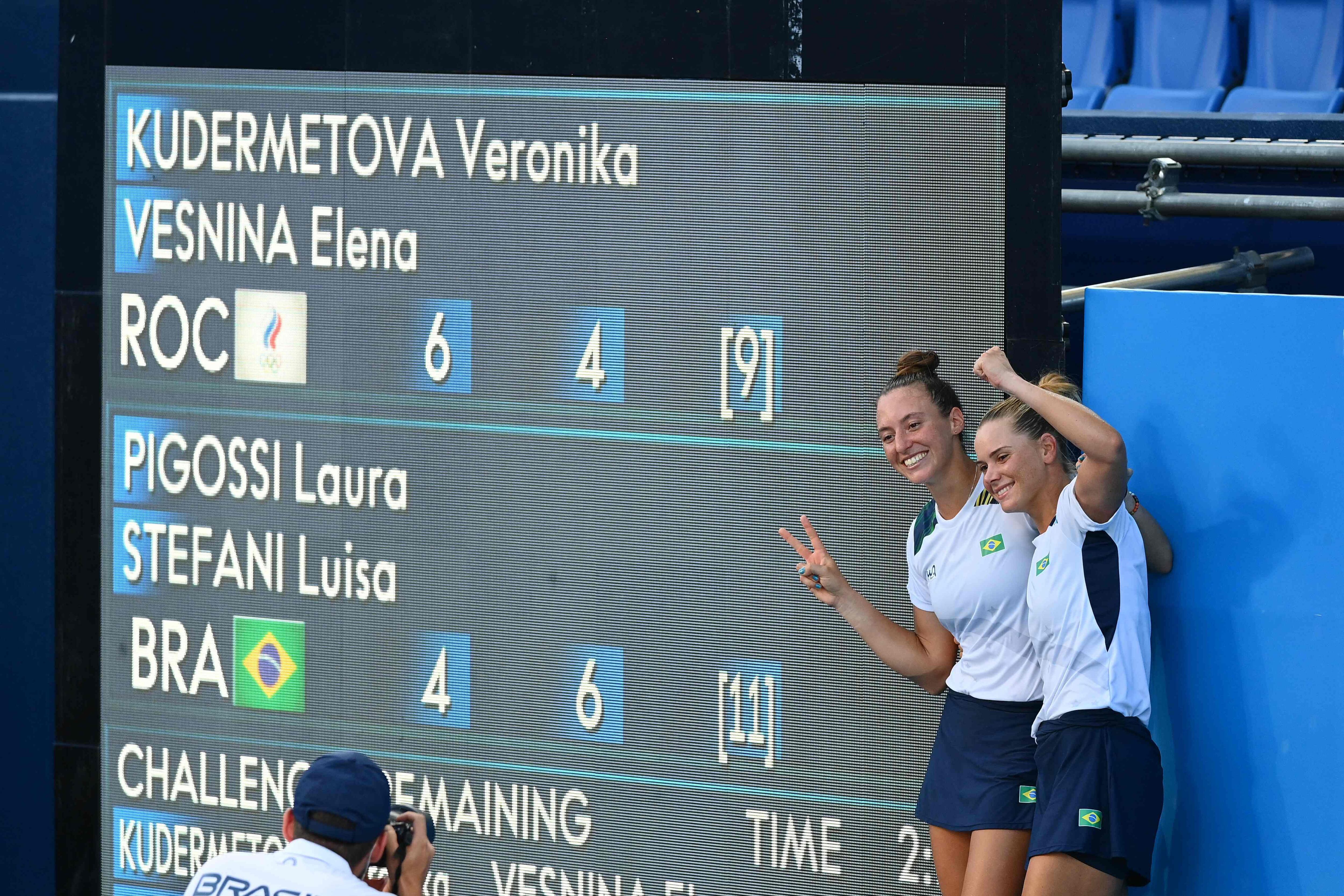 Laura Pigossi (à direita) e Luisa Stefani posam ao lado do quadro de resultados após conquistarem o bronze olímpico inédito.