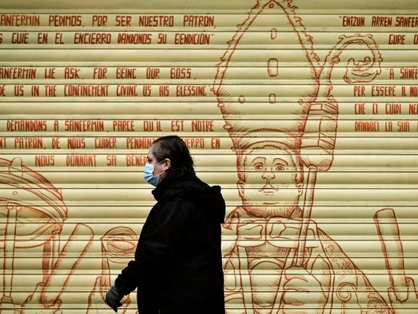 A woman wearing a face mask walks in front of graffiti depicting Saint Fermin, with writing in many different languages asking for blessings, in Pamplona, northern Spain, Wednesday, March 18, 2020. Spain will mobilize 200 billion euros or the equivalent to one fifth of the country's annual output in loans, credit guarantees and subsidies for workers and vulnerable citizens, Prime Minister Pedro Sanchez announced Tuesday. For most people, the new coronavirus causes only mild or moderate symptoms. For some, it can cause more severe illness, especially in older adults and people with existing health problems. (AP Photo/Alvaro Barrientos)