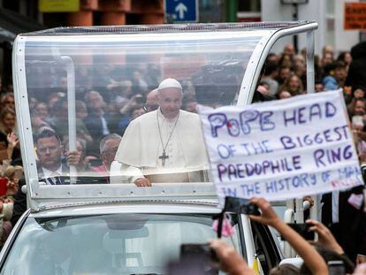 Ativista mostra um cartaz contra o papa Francisco (c), que percorre as ruas do centro de Dublin a bordo do papamóvel, no sábado. No vídeo, o Papa reza em Dublin pelas vítimas dos abusos.