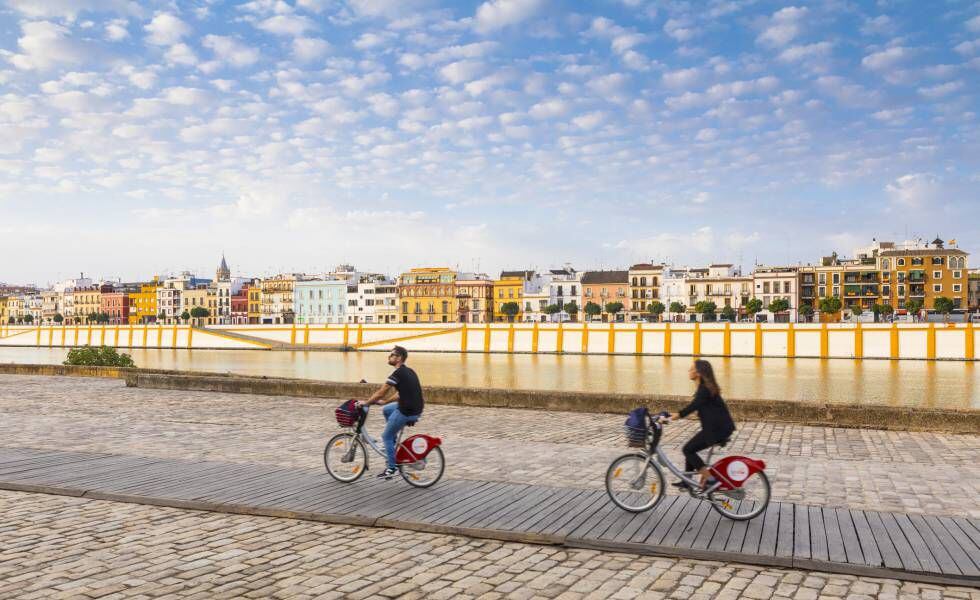 Dois turistas em bicicletas públicas de aluguel pedalando ao longo do rio Guadalquivir, em Sevilha, com o bairro de Triana ao fundo.