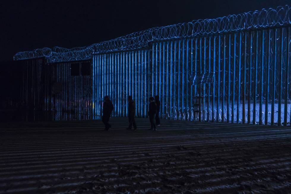 Um grupo de pessoas caminha diante do muro fronteiriço de Tijuana, na sexta-feira à noite.