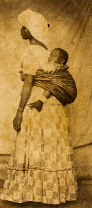 Mulher negra posando em estúdio com traje comum aos escravos brasileiros do séc. XIX: pano-da-costa utilizado para carregar criança, turbante, saia comprida.