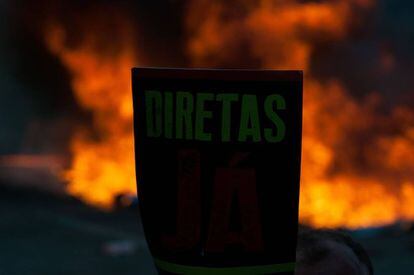 Manifestante com um cartaz em defesa de eleições antecipadas diretas, em Brasília. Ao fundo, barricada com fogo na Esplanada dos Ministérios.