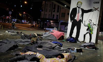 Moradores de rua descansam em frente a um grafite representando Bolsonaro e Trump.