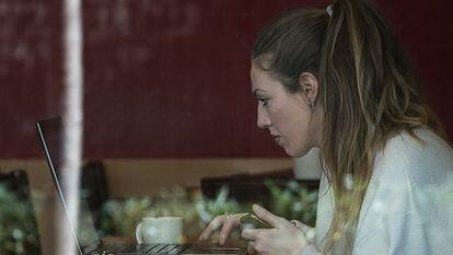 Una joven consulta su dispositivo móvil y su portátil a la vez mientras desayuna en el interior de una cafetería.