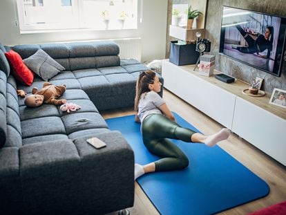 Uma mãe faz exercício diante da televisão enquanto seu bebê descansa no sofá.