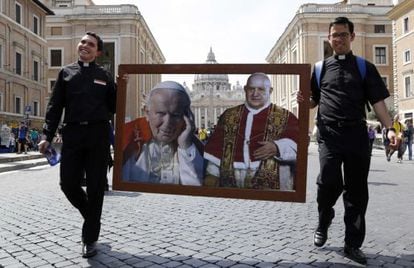 Dois sacerdotes caminham pela Praça de San Pedro com um quadro com os dois papas que serão canonizados no próximo dia 27.