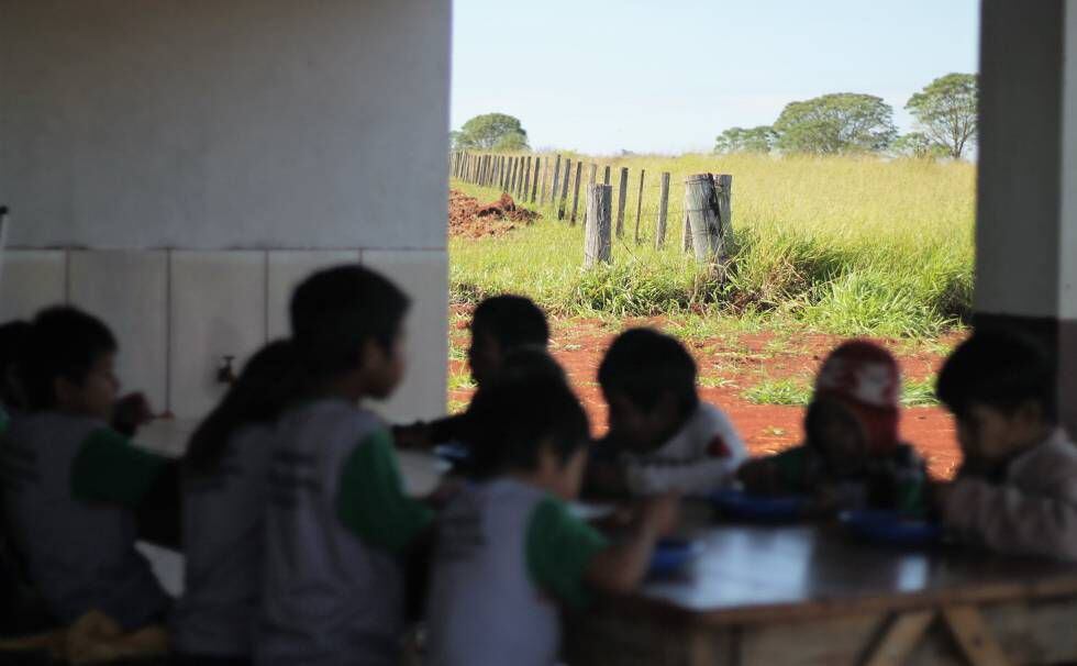 Estudantes da escola indígena de Guyraroká comem a merenda no intervalo entre as aulas. Ao fundo, o limite entre a comunidade e área rural vizinha.